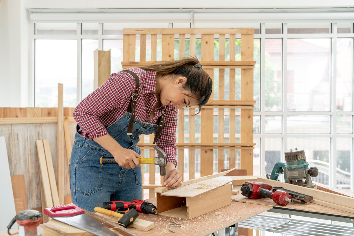 A Confident female carpenter uses a hammer and nail to hammering the wooden bar at the carpentry shop. Asian handywoman apprentice working in a workshop. DIY woodworking crafts and Hobby concepts.